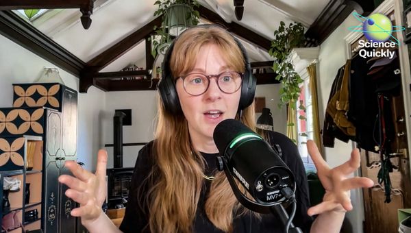A woman in glasses speaking into a microphone with hands raised