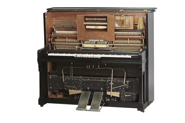 The exposed internal pneumatic mechanisms, keyboard, foot pedals, and central perforated paper roll of a vintage player piano isolated on a white background.