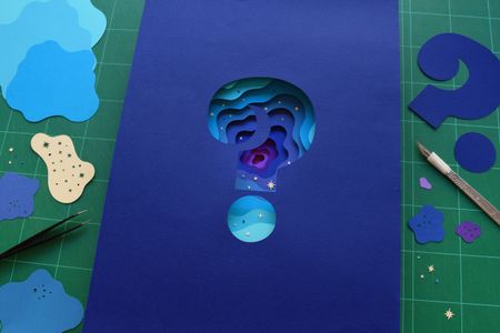 Question mark-shaped cut out revealing layers of cut paper underneath in graduating colors of blue and purple with tweezers and an xacto tool on the sides.