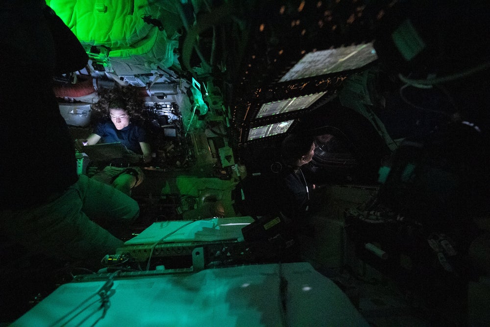 Una vista desordenada dentro de una nave espacial a oscuras, con una mujer cuyo rostro está iluminado por una pantalla de computadora.