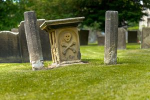 An old grave marker in a cemetary shows a skull and crossed bones.