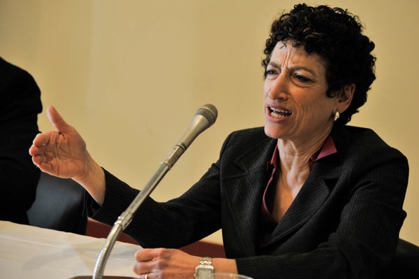Naomi Oreskes in a black suit, sitting down and speaking into a microphone.