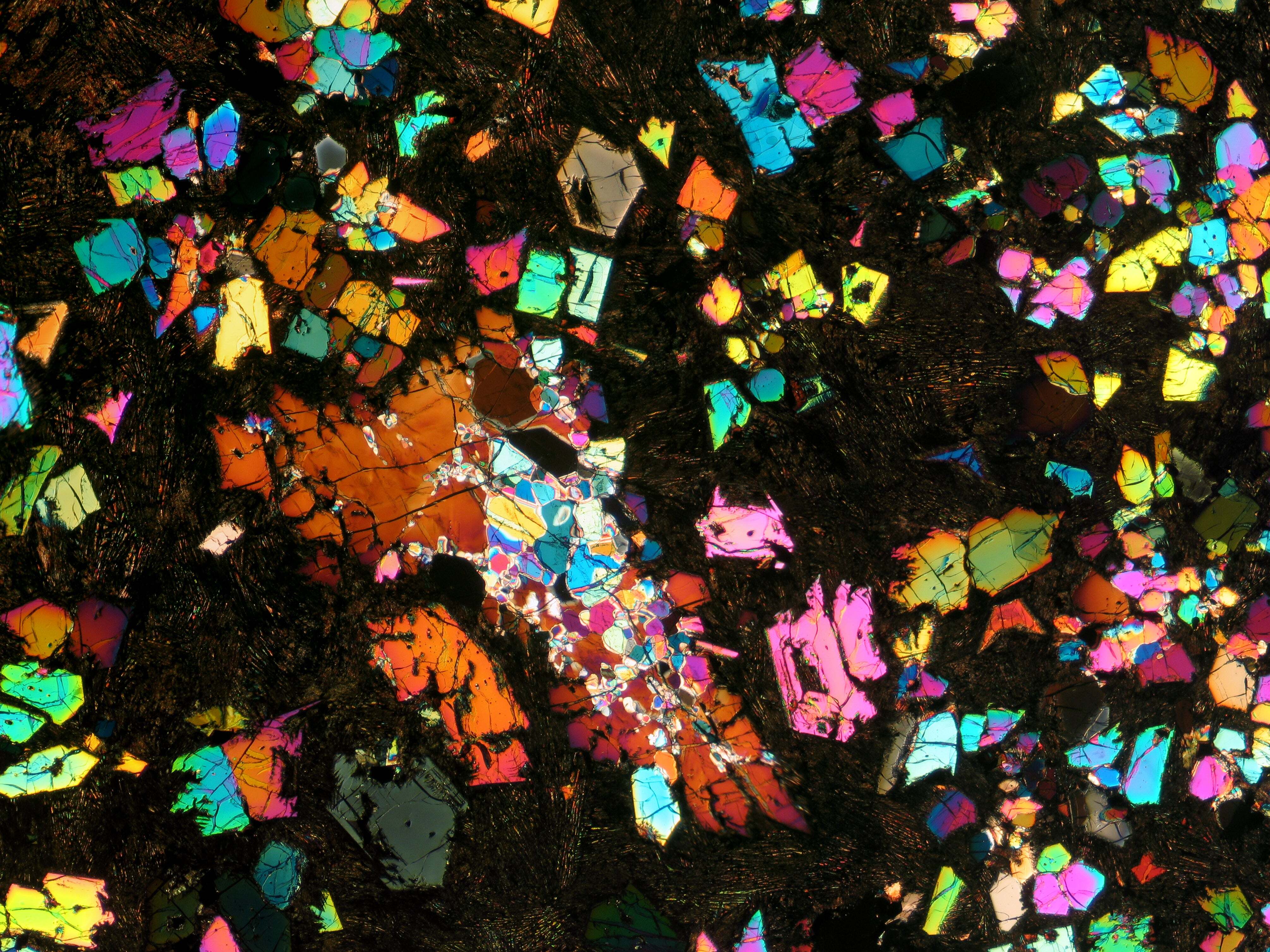 A thin section of a meteorite dotted with rainbowlike, oddly shaped mineral flecks within a matrix of darker rock.