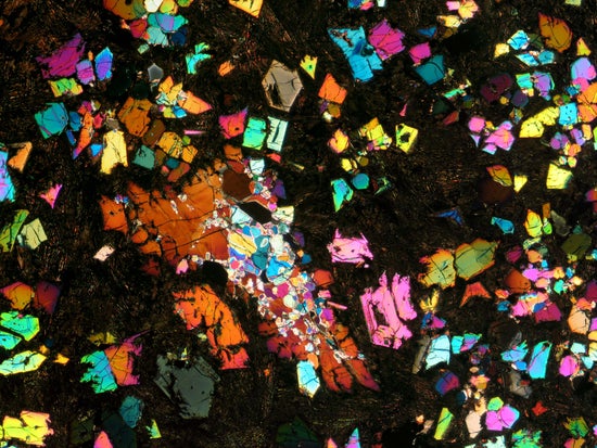 A thin section of a meteorite dotted with rainbowlike, oddly shaped mineral flecks within a matrix of darker rock.