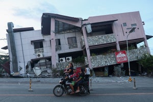 Several people on a motorcycle in front of a large building that has sunk down at a weird angle and shows lots of damage.