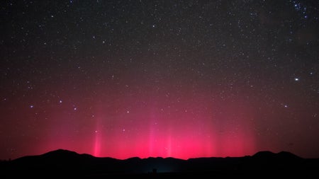 Rippling red light emerges into a starry night sky from behind a hilly silhouette.