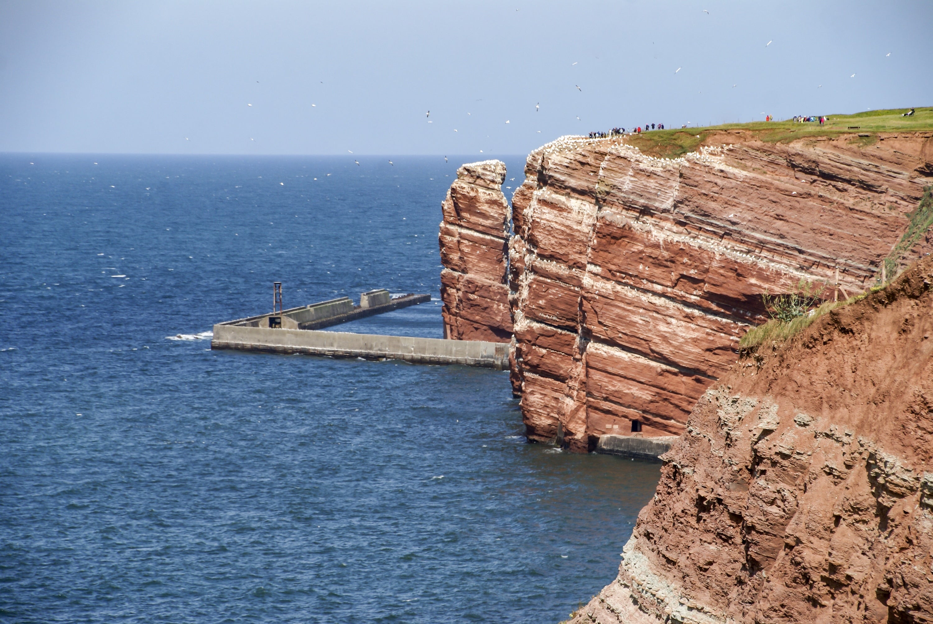 An angular set of cliffs juts into the sea, tiny people just visible along its top edge