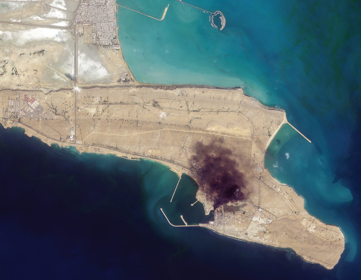 Overhead satellite view of a narrow coastal naval base, with a large dark area of apparent strike damage near the harbor.