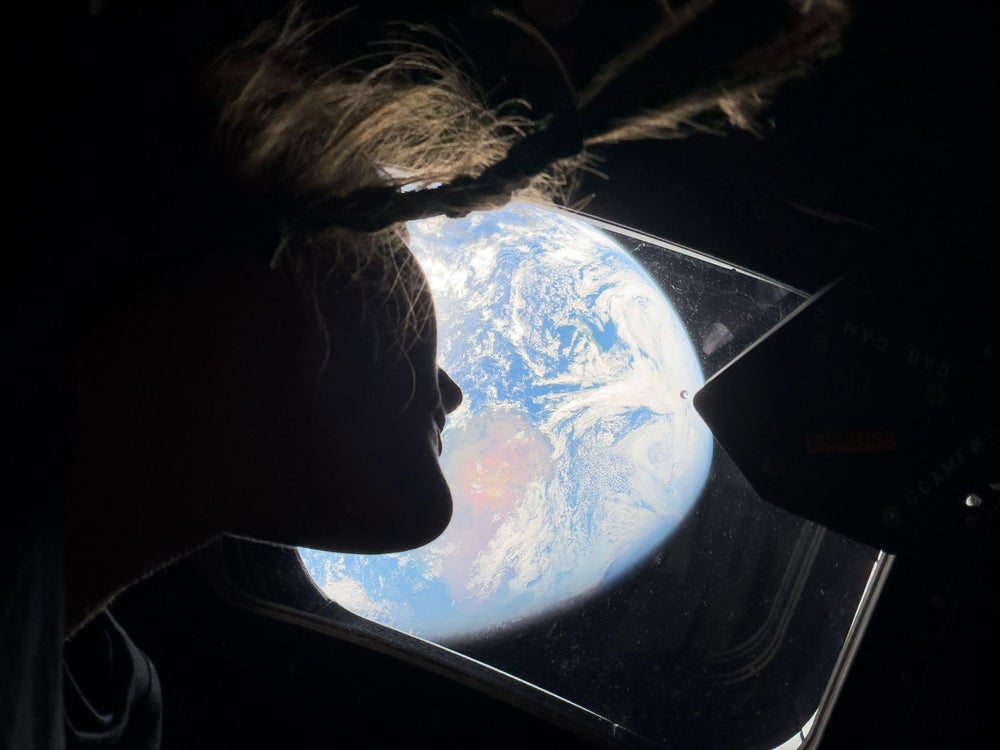 La astronauta Christina Koch mira por la ventana de la nave espacial Orion con una Tierra de color blanco pálido a lo lejos.