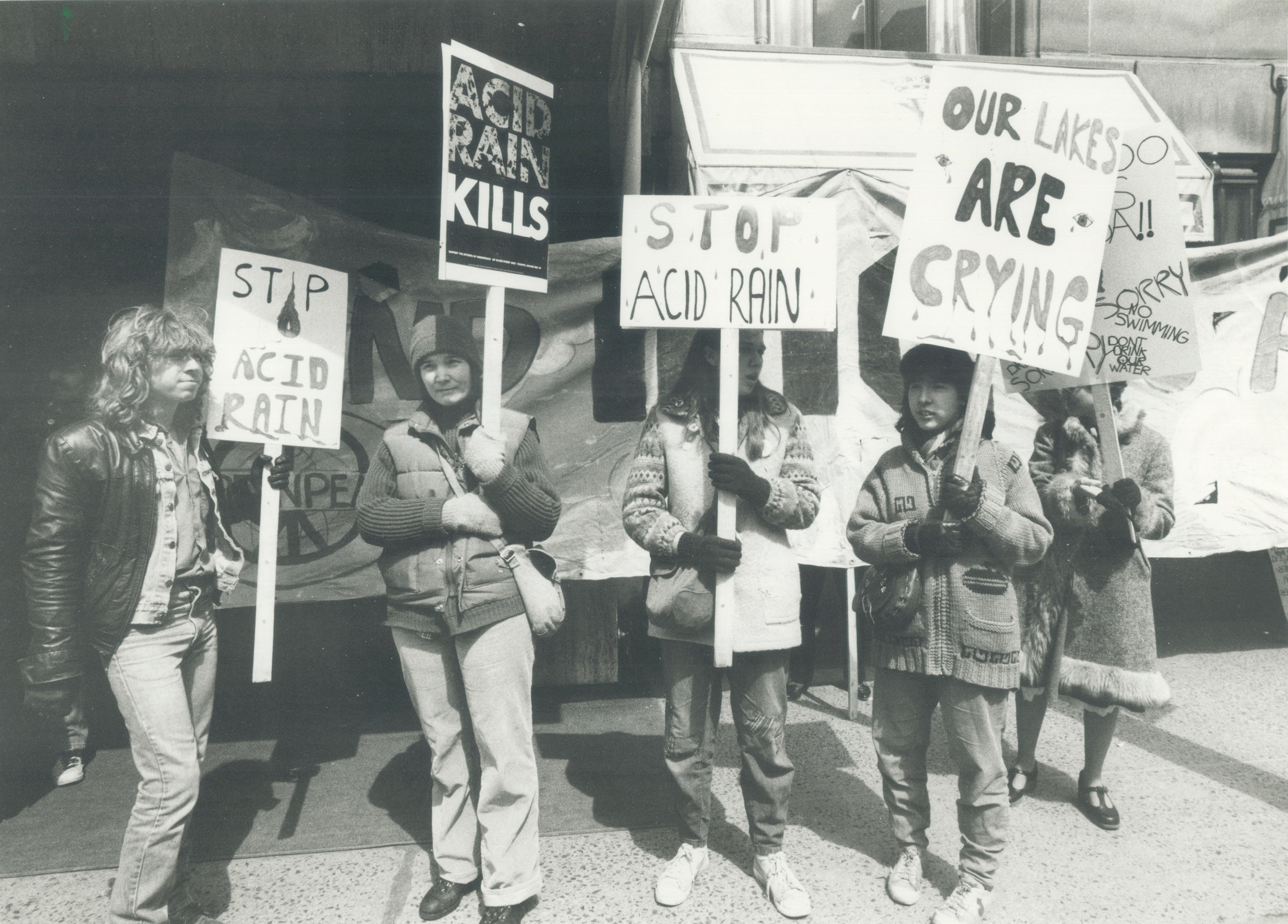 A black-and-white photo showing a group of give people holding signs with phrases like "Stop Acid Rain," "Acid Rain Kills" and "Our Lakes Are Crying". They are standing in front of a larger banner.
