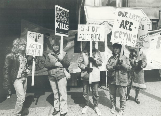 A black-and-white photo showing a group of give people holding signs with phrases like "Stop Acid Rain," "Acid Rain Kills" and "Our Lakes Are Crying". They are standing in front of a larger banner.