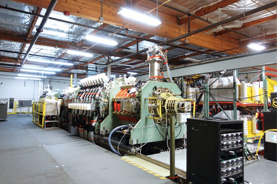 A long, pale green fusion prototype stretches across a bright industrial facility, with metal chambers, cables and control equipment along its length.