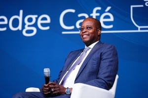 Wayne Frederick sitting in a blue suit and holding a microphone, against a blue background.