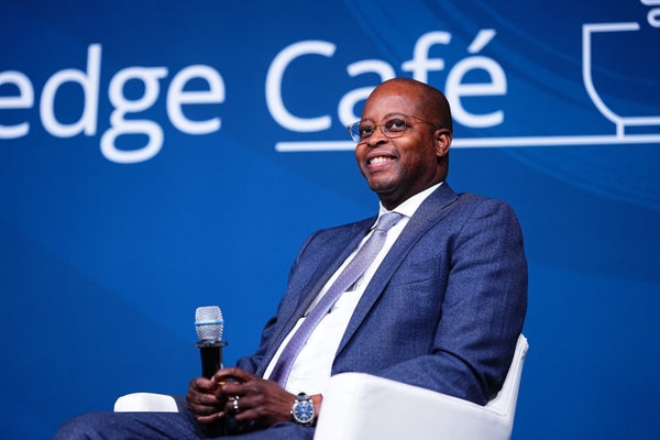 Wayne Frederick sitting in a blue suit and holding a microphone, against a blue background.