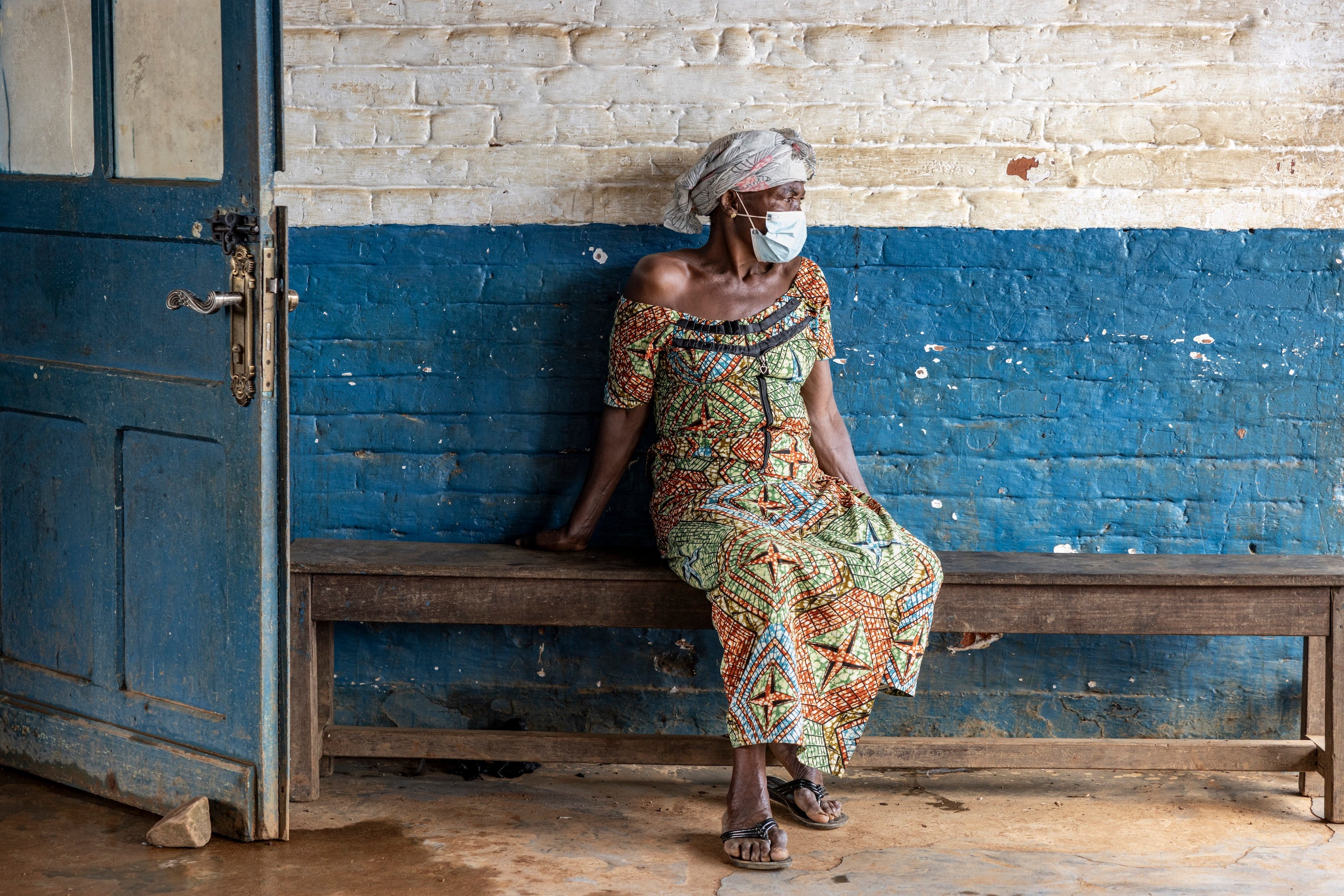A woman in a colorful dress and head wrap wearing a surgical mask sits on a bench in frnot of a blue and white brick wall, looking to the side.