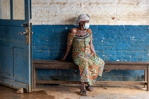 A woman in a colorful dress and head wrap wearing a surgical mask sits on a bench in frnot of a blue and white brick wall, looking to the side.