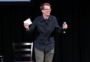 A smiling speaker stands onstage holding a microphone in one hand and a small white card in the other.