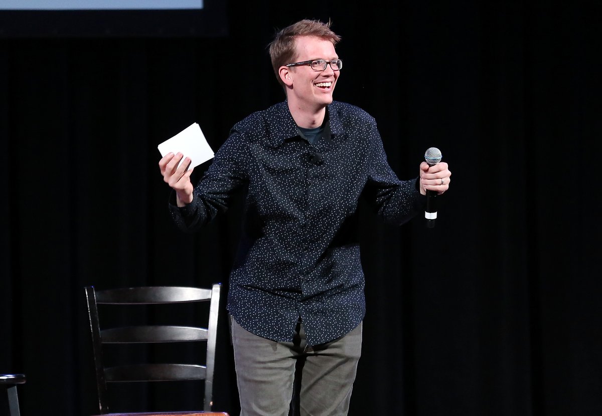 A smiling speaker stands onstage holding a microphone in one hand and a small white card in the other.