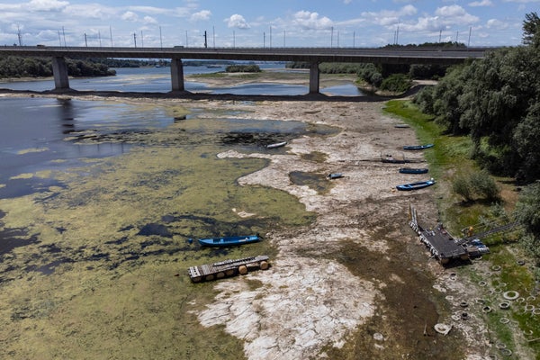 Boats site on a dried river bed with tan soil visible the water reduced to a much narrower channel in the middle of the riverbed. A bridge is seen in the background.