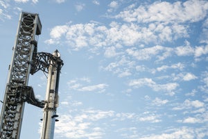 A tall stainless-steel rocket stands beside a launch tower under a blue sky with scattered clouds.