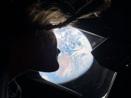 Photograph of astronaut Christina Koch looking out window of Orion spacecraft at a distant Earth