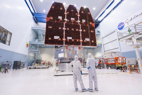 Two bunnysuited engineers stand facing away from the camera in a large clean room at NASA's Goddard Space Flight Center, gazing at flight hardware for the space agency's Nancy Grace Roman Space Telescope.