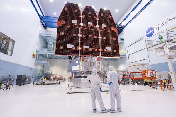 Two bunnysuited engineers stand facing away from the camera in a large clean room at NASA's Goddard Space Flight Center, gazing at flight hardware for the space agency's Nancy Grace Roman Space Telescope.