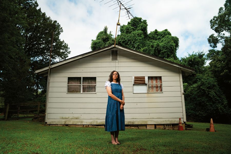 A woman in a long blue dress stands on the lawn in front of a small one-story house shaded by trees.
