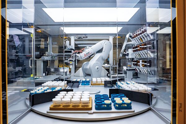 A large, white robotic arm works inside a glass enclosure, surrounded by trays of capped sample jars.