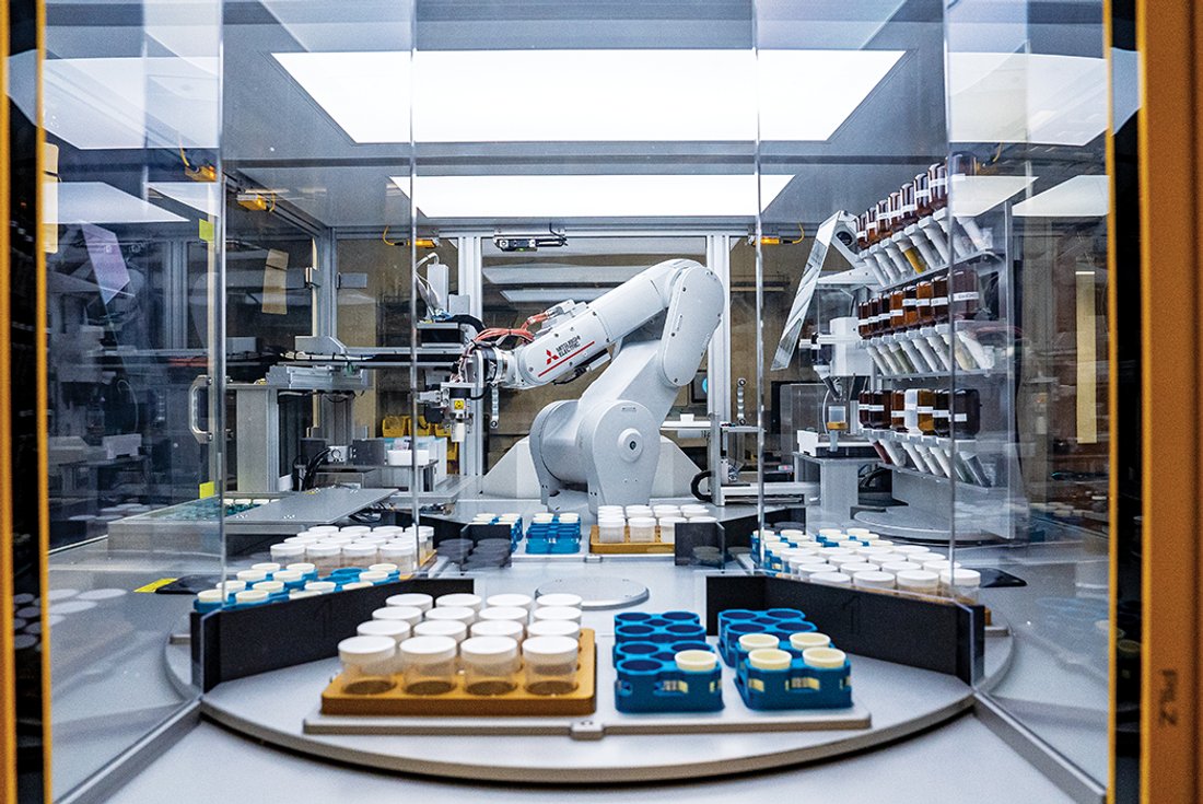 A large, white robotic arm works inside a glass enclosure, surrounded by trays of capped sample jars.