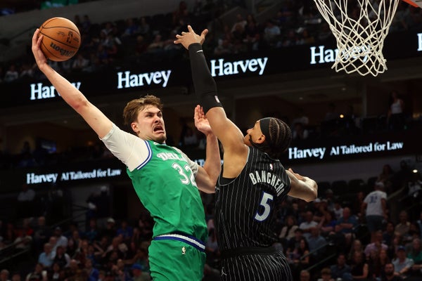 A basketball player in a green jersey that displays “Dallas” and the number “32” tries to dunk the ball while a player in a black jersey that displays the number “5” tries to defend the basket.
