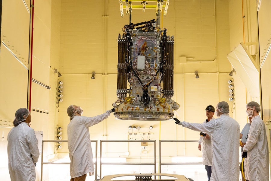 A spacecraft covered foil hangs above engineers in white uniforms in a large testing chamber.