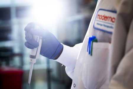 This closeup of a researcher in a lab shows the person’s gloved hand with a pipette device and a blurry background. The researcher is wearing a white lab coat with a Moderna patch on the front.