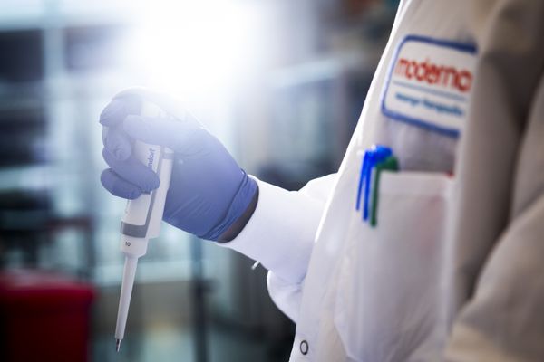 This closeup of a researcher in a lab shows the person’s gloved hand with a pipette device and a blurry background. The researcher is wearing a white lab coat with a Moderna patch on the front.