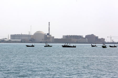 The low, gray buildings of a power plant seen with Persian Gulf waters and boats in the foreground