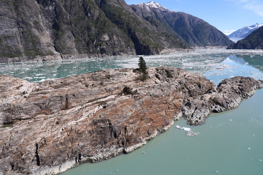 A nearly barren rocky island, surrounded by teal waters. Green cliffs behind it have bare spo