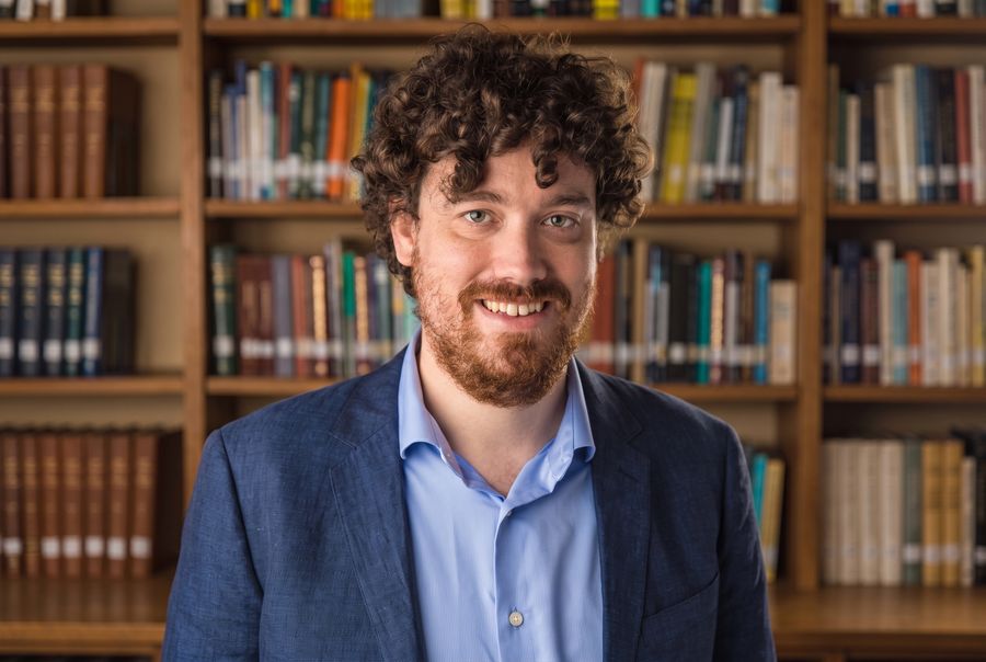 Headshot of a curly-haired mathematician smiling in front of a stacked booshelf.