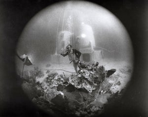View through a rounded hole of a diver in front of two cylinders of underwater habitat on the seafloor.