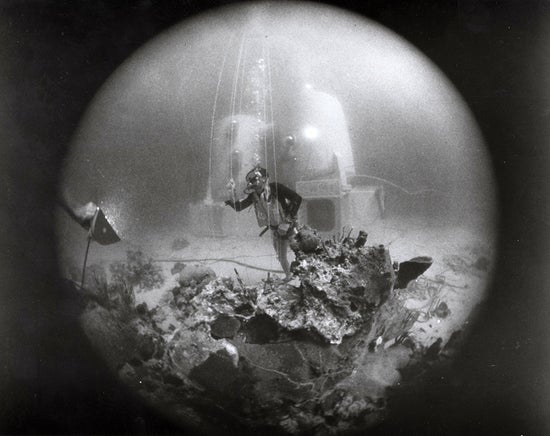 View through a rounded hole of a diver in front of two cylinders of underwater habitat on the seafloor.