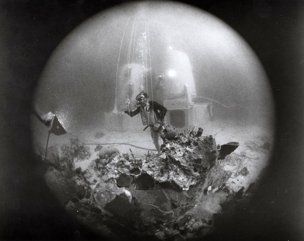 View through a rounded hole of a diver in front of two cylinders of underwater habitat on the seafloor.