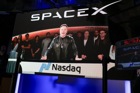 Elon Musk is shown on a giant screen with the SpaceX logo in the background. The screen is displayed at the Nasdaq MarketSite in New York.