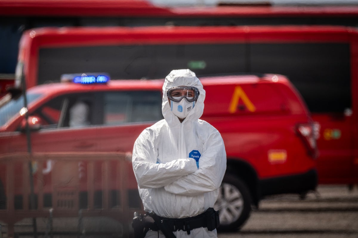 A person in a hazmat suit and mask stands with their arms folded in front of multiple emergency vehicles.