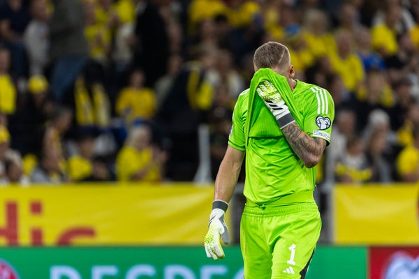 A goalkeeper in neon green kit wipes his face with his shirt. The crowd and signs are visible in the background.