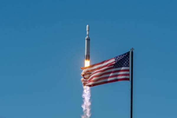 A SpaceX Falcon Heavy rocket launches into a clear blue sky, with a billowing American flag in the foreground.