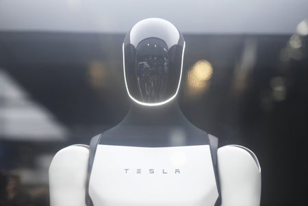 A sleek white humanoid robot with a glossy black visor and illuminated facial outline, branded with “TESLA,” stands against a softly blurred background.