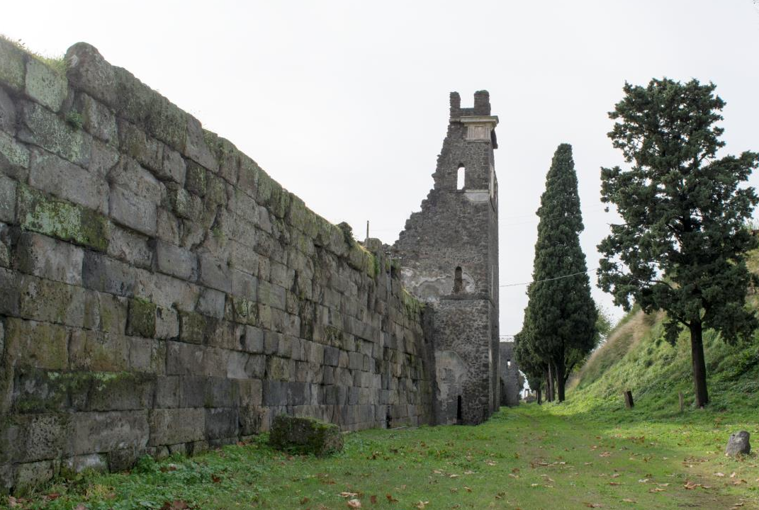 A stone wall with a partially destroyed tower seen at an angle with two tall trees next to it