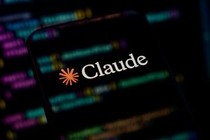 The Claude by Anthropic logo is displayed on a mobile phone screen placed over a computer monitor showing lines of programming code in Python and React.