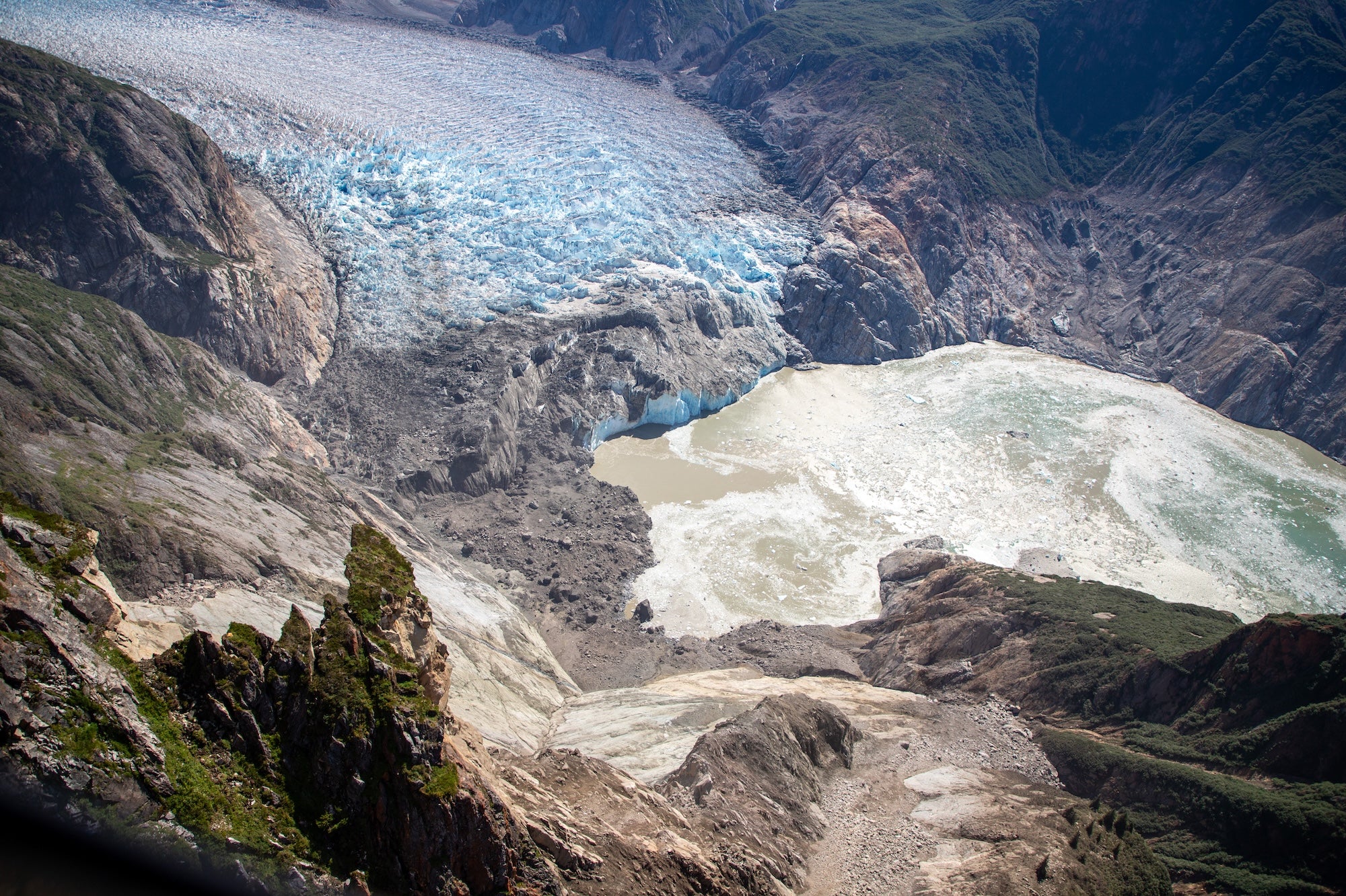 An aerial view of the end of a fjord, with the white-blue ice of a glacier at the top of the photo, surrounded by rocky walls, some of which have collapsed, leaving a swirl of muddy water
