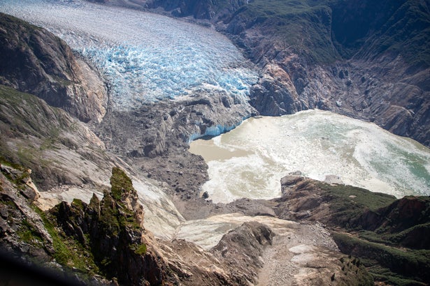 An aerial view of the end of a fjord, with the white-blue ice of a glacier at the top of the photo, surrounded by rocky walls, some of which have collapsed, leaving a swirl of muddy water