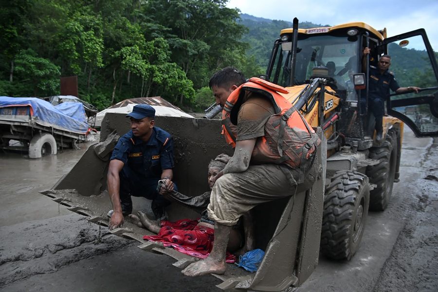 Two rescue personnel sit inside the scoop of an excavator with a person covered in mud.